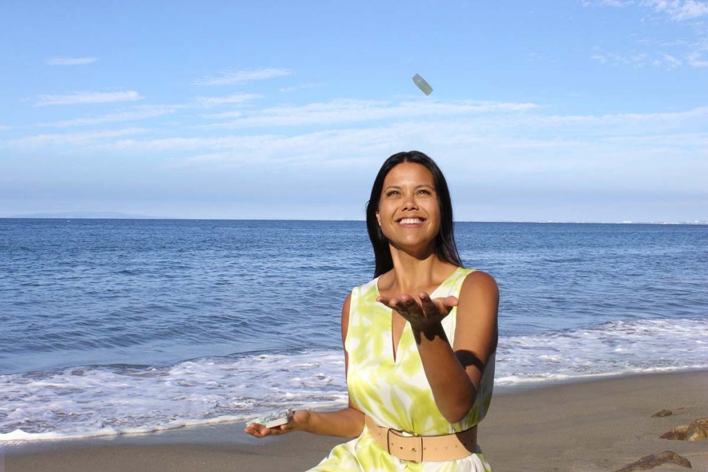 Tiffany Buzzano, the founder of eco-friendly small skincare company, Dew Mighty, walking on the beach in a yellow dress tossing one of her solid serum bars in the air.