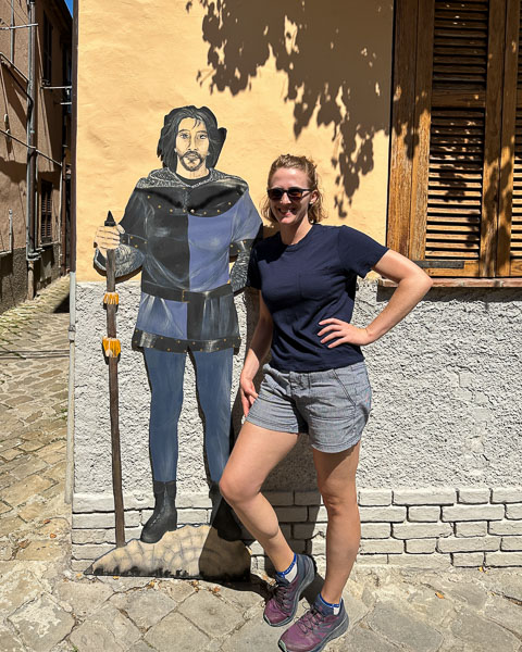 The review author wearing a navy blue Unbound Merino wool t-shirt while traveling in Italy, standing next to a medieval knight cutout in Fabriano, Italy. ©KettiWilhelm2023