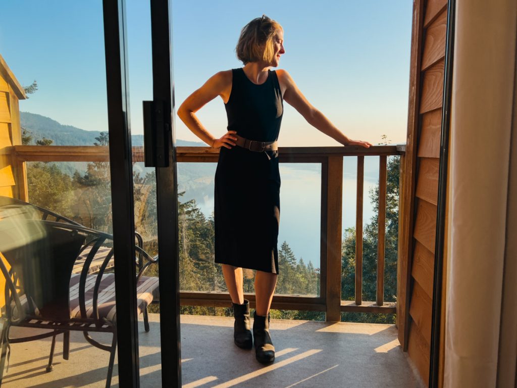 The blogger/reviewer, a white woman with short blond hair, wearing a sleeveless, black merino wool travel dress on a balcony in front of a mountain view on Vancouver Island. ©KettiWIlhelm2022