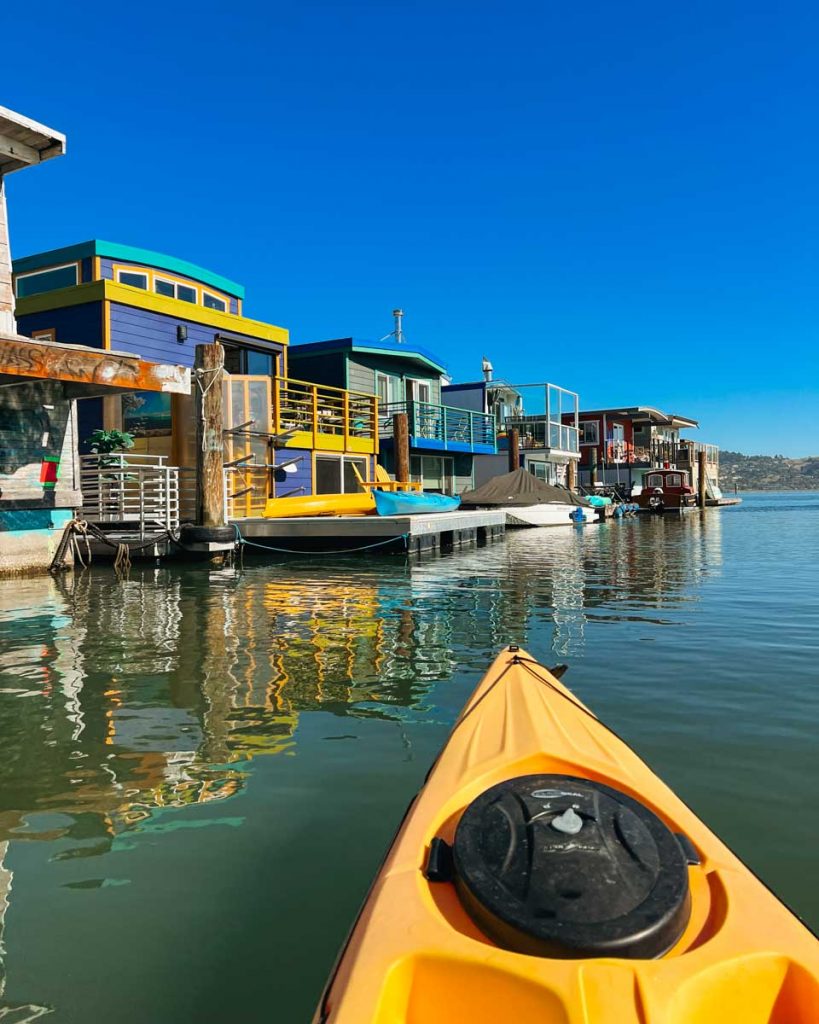 The front of a bright yellow kayak pointing toward the colorful floating houses of Sausalito, near San Francisco. ©KettiWilhelm2022