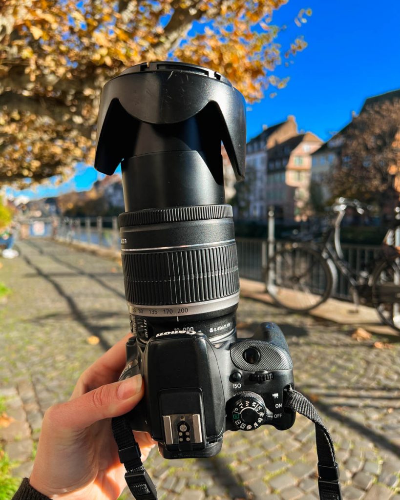 Holding up my Canon DSLR camera, with the zoom lens crooked after I was hit by a car in France, in front of the river walk in Strasbourg. ©KettiWilhelm2023