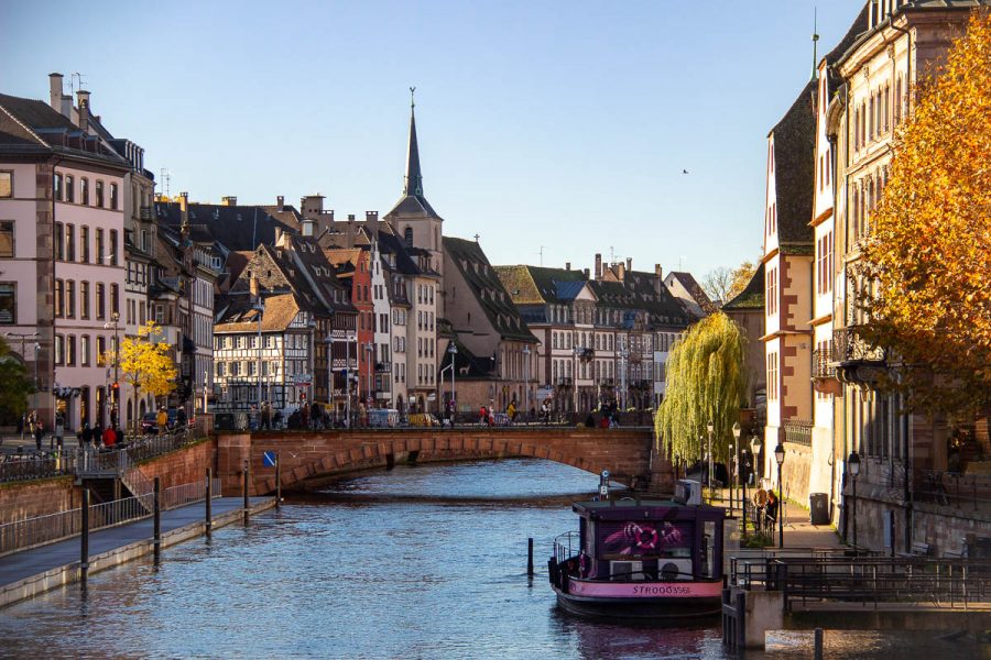 The Ill River traverses Strasbourg, France, under stone bridges, with sunlight filtering through the old buildings, and river cruise boats for tourists anchored at the shore. ©KettiWilhelm2023