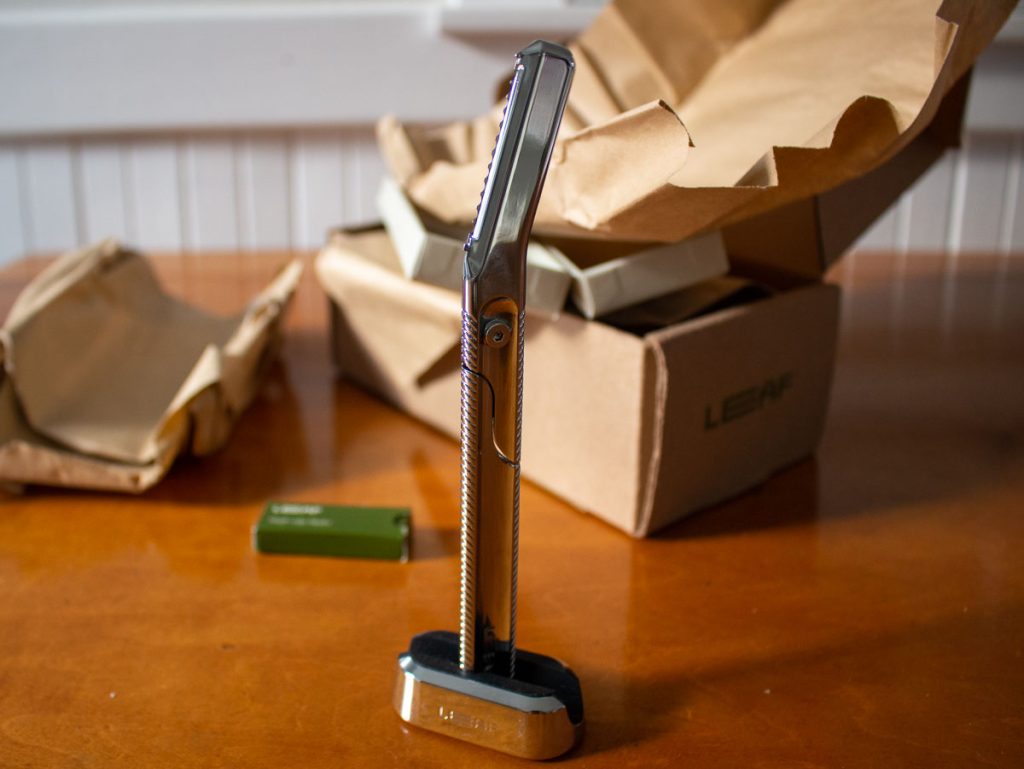 Leaf Shave's metal dermaplaner tool, reviewed in this article, in its stand, both in the color silver, with Leaf's plastic-free packaging sitting behind it on a wooden counter. ©KettiWilhelm2023