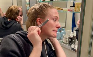 Learning how to dermaplane at home for the first time – the review writer leans toward a mirror with a reusable Leaf Shave Co. brand dermaplaning tool in her hand, with a nervous, grimacing expression. ©KettiWilhelm2023