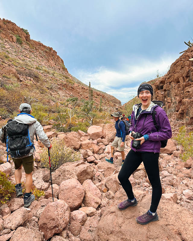 Ketti, the travel writer behind this blog, grinning in a desert arroyo, and modeling the dress code for UnCruise in Baja: Casual with light layers. ©KettiWilhelm2023