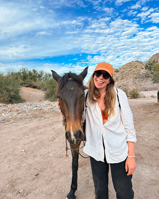 My Baja travel buddy posing with a burro after our desert burro rides, wearing a white linen shirt, linen pants, and a baseball cap. ©KettiWilhelm2023