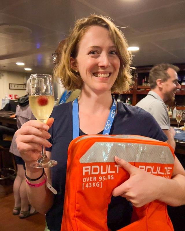 Ketti, the traveler writing this review, wearing a bright orange life jacket and grinning with a glass of champagne in her hand during the safety meeting on the first night of the Baja UnCruise trip. ©KettiWilhelm2023