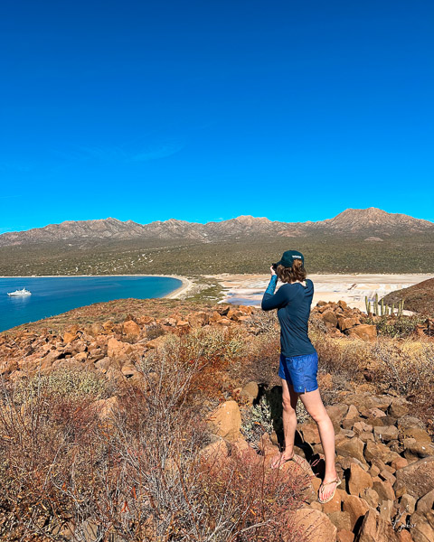 The writer looking down on turquoise waters with a small ship in the bay in front of an empty white sand beach on an island in Baja California Sur. ©KettiWilhelm2023