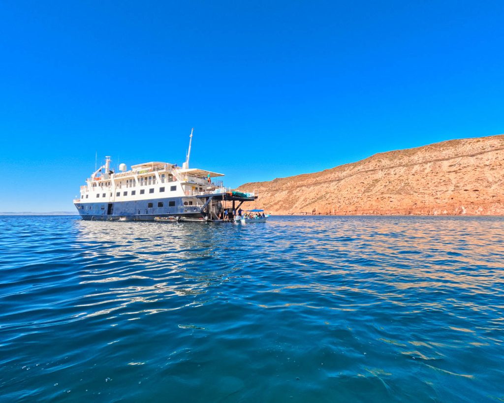 UnCruise’s small cruise ship, the Safari Voyager, in the Sea of Cortez, Baja California, seen over the water from a skiff with the desert mountain behind. ©KettiWilhelm2023