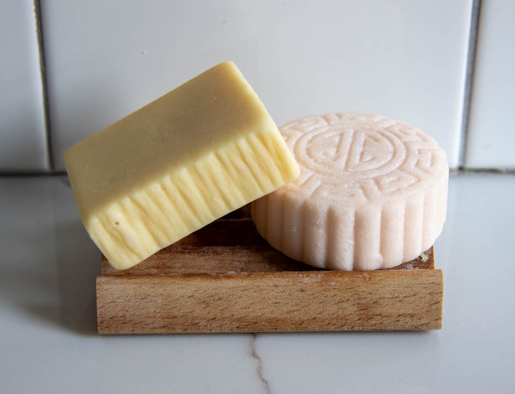 My Viori conditioner bar rests on top of my shampoo bar, both reviewed in this blog post, on a wooden soap dish in front of a white tile shower. ©KettiWilhelm2023
