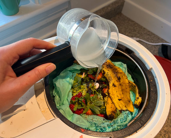 Pouring an ounce of two of water into the bucket of the Lomi, over a LomiPod and food scraps, including a banana peel. ©KettiWilhelm2023