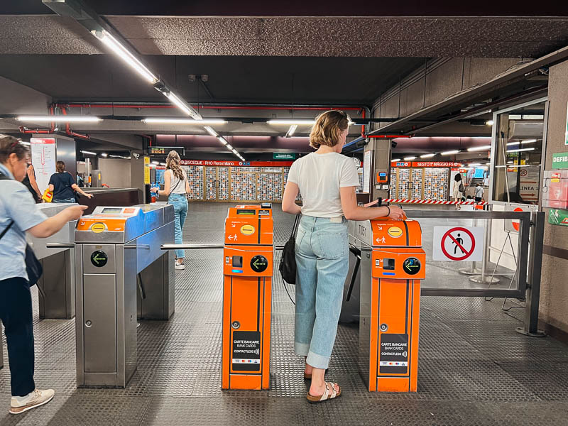 Travel blogger Ketti Wilhelm swiping her credit card to enter the subway system in Milan, Italy, between two orange metro turnstiles. ©KettiWilhelm2023