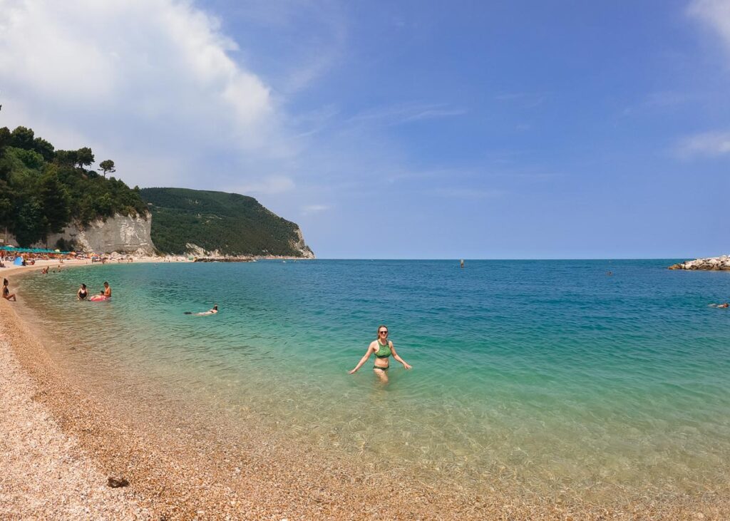 The author and travel writer, a white woman, standing in the turquoise water of the Adriatic Sea, on a beach in Sirolo at the end of our Marche by bike e-bike tour in Italy. ©KettiWilhelm2023