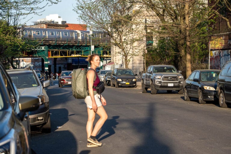 Travel blogger walking across a street in New York City wearing a travel backpack carrying everything she's packing for weeks of travel packing light in Europe. ©KettiWilhelm2026