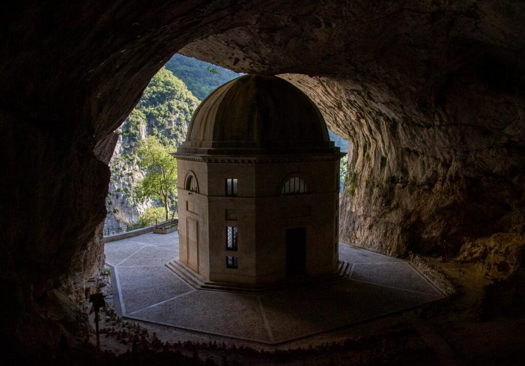 Il Santuario della Madonna di Frasassi (the Sanctuary of the Madonna of Frasassi), an ancient chapel carved into a cave in Marche, Italy, with the green of the hillside showing behind it. ©KettiWilhelm2023
