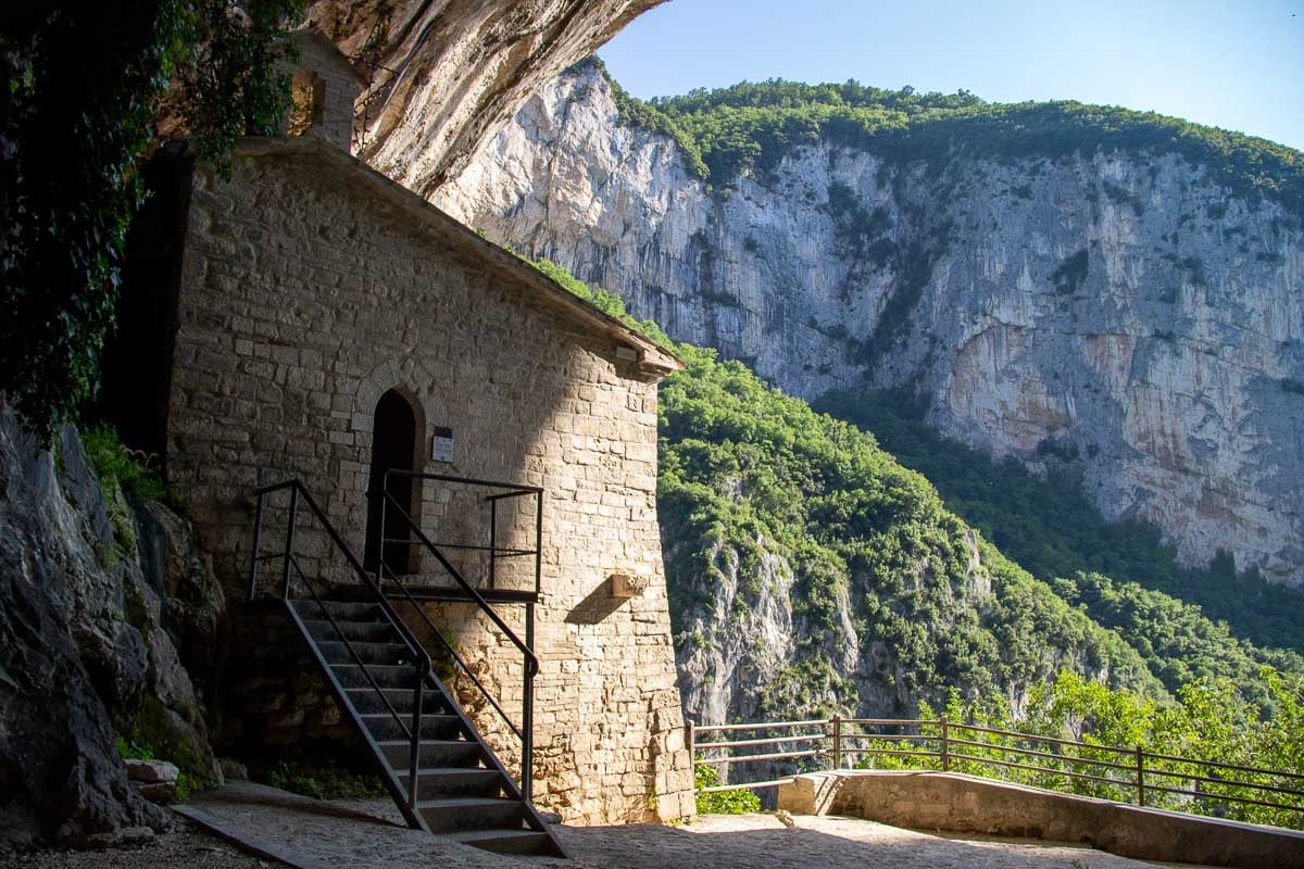 Il Santuario della Madonna di Frasassi (the Sanctuary of the Madonna of Frasassi), an ancient chapel carved into a cave in Marche, Italy, with the green of the hillside showing behind it. ©KettiWilhelm2023