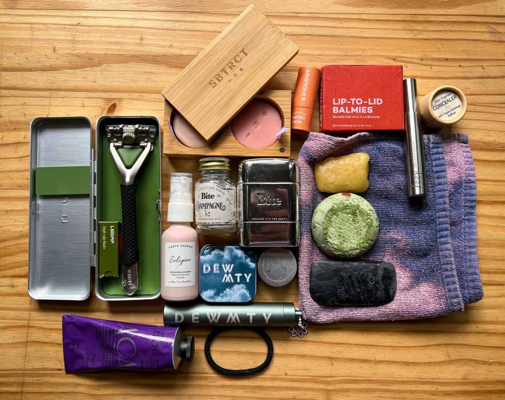 My entire toiletry kit that helps me pack light: solid toothpaste tablets in a small glass jar, a plastic-free razor in a metal carrying case, travel-sized moisturizer in an aluminum tube, shampoo bar, conditioner bar, and face wash bar, solid skincare serum, and assorted sustainable cosmetics. ©KettiWilhelm2023