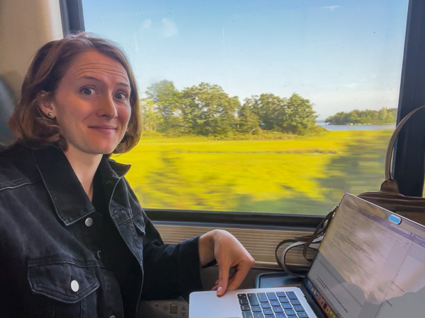 Ketti Wilhelm, the author of this guide to packing light, sitting on a train with a green field in the background, writing about packing light on her laptop. ©KettiWilhelm2023