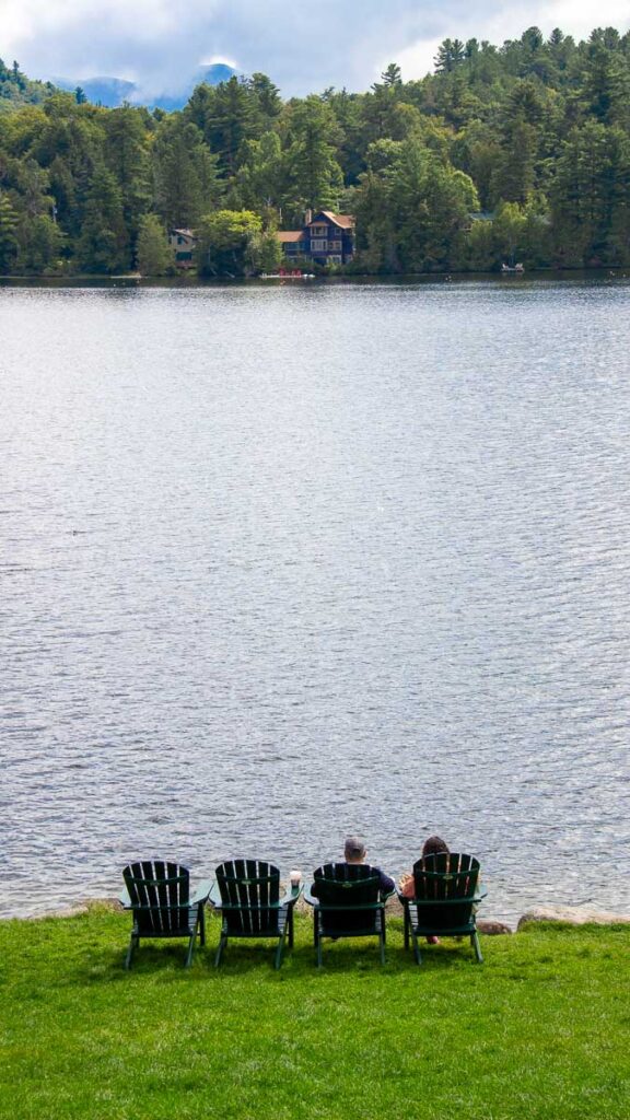 Four Adirondack chairs sit facing Mirror Lake, in Lake Placid, New York, with a backdrop of the Adirondack High Peaks behind. ©KettiWilhelm2023