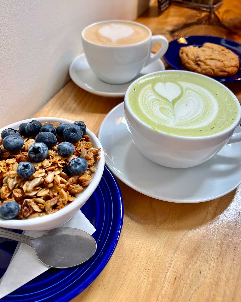 A bowl of granola with fresh blueberries, a matcha latte with oat milk, and an oat milk latte, all in eco-friendly reusable ceramic cups at Origin Coffee Co – the best coffee shop I found in Lake Placid, in the Adirondacks. ©KettiWilhelm2023