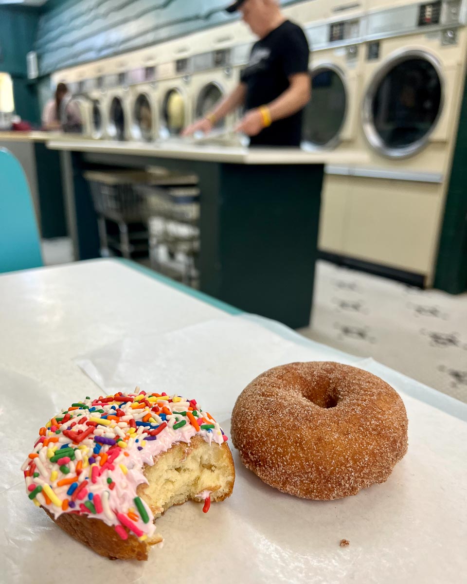 Two donuts on a piece of paper on the table of a booth at the Donut Shoppe in Tupper Lake, NY, with laundry machines behind. ©KettiWilhelm2023