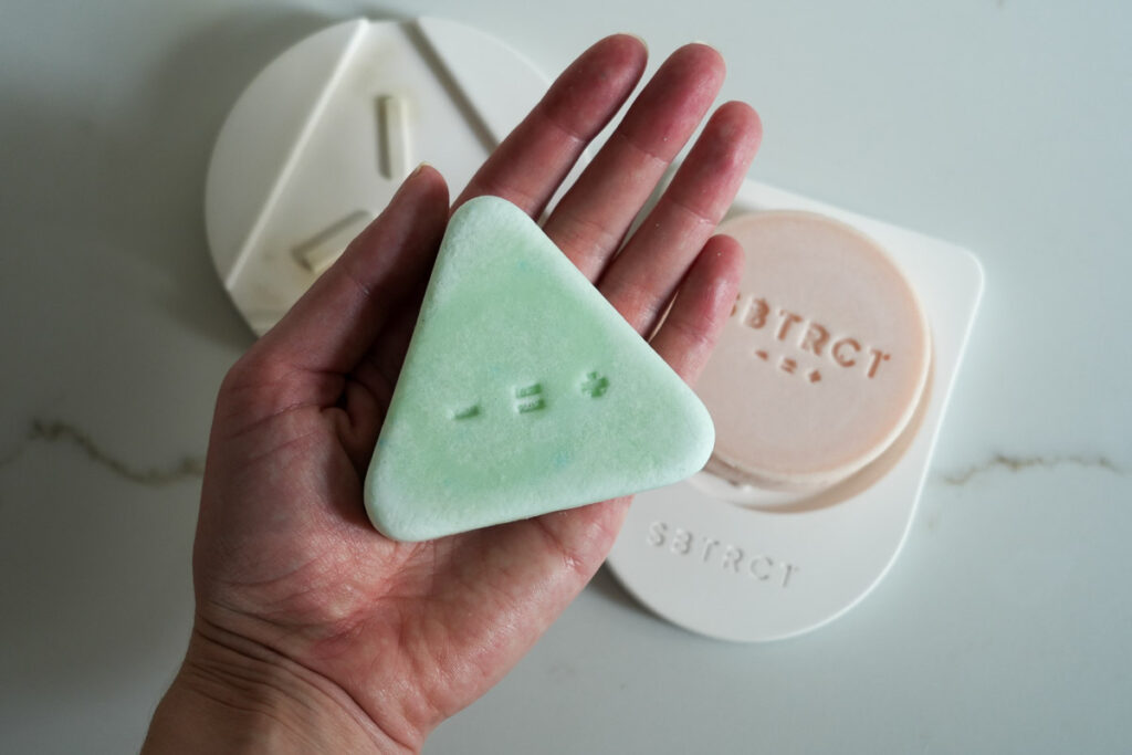 A bar of solid Clarifying Facial Exfoliator from SBTRCT skincare that's the size of my entire palm, where it's sitting, with their Gentle Foaming Cleanser and diatomite soap dishes in the background. ©KettiWilhelm2023