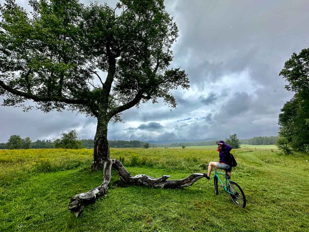 The author of this Adirondack guide sitting on her bike in the rain next to a tree at Heaven Hill Trails, an easy hike and biking area in the Adirondacks. ©KettiWilhelm2023