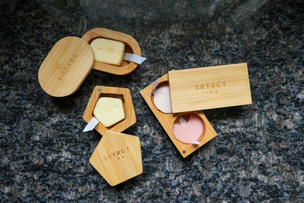 All three of my bamboo travel containers for SBTRCT's plastic-free skincare line, sitting open, showing the solid skincare products inside, on a black marble bathroom counter. ©KettiWilhelm2023