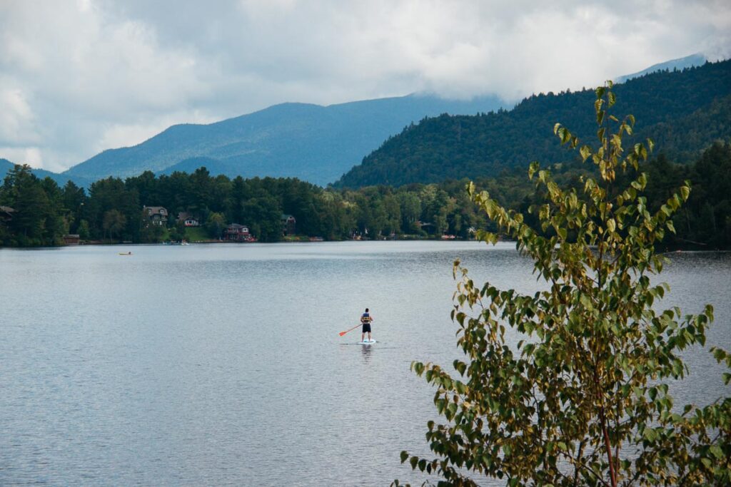 From the balcony of the Golden Arrow Lakeside Resort, a sustainable hotel in the Adirondacks, a solo man is seen on a stand-up paddle board (SUP) in the middle of Mirror Lake. ©KettiWilhelm2023