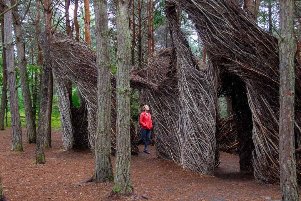 The author of this Adirondack weekend getaway itinerary, in an orange jacket, walking through the Hopscotch stick art sculpture in the forest at the Wild Center, a fun activity in the Adirondacks. ©KettiWilhelm2023