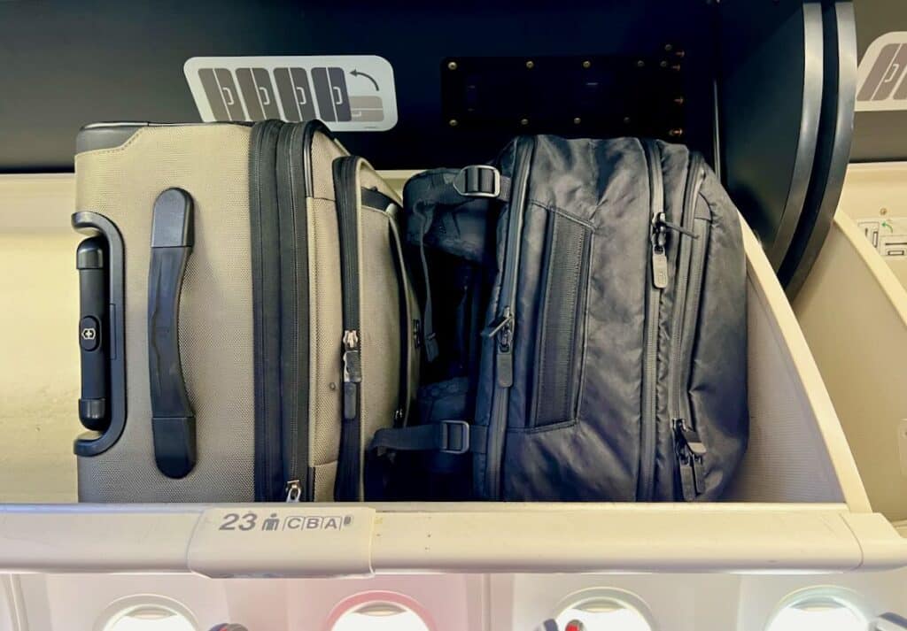 Comparing a travel backpack vs luggage as a carry on, with a max carry-on size suitcase and Tortuga backpack next to each on an airplane's overhead luggage rack. ©KettiWilhelm2023