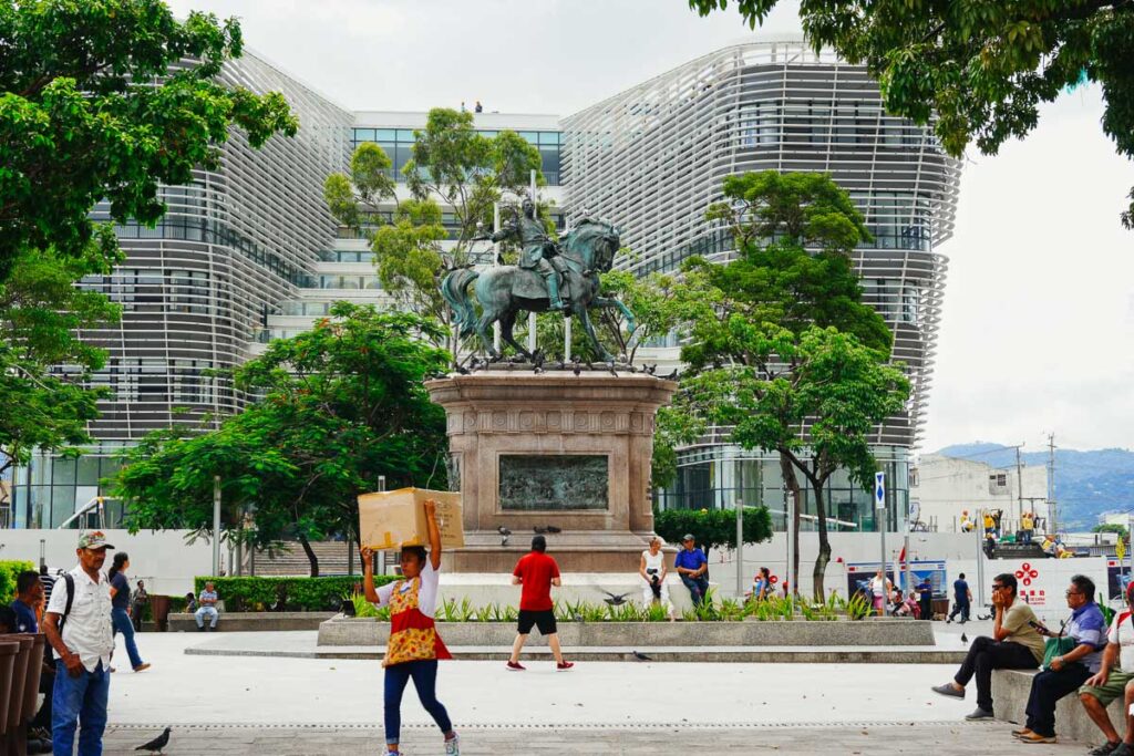 A woman carries a large box on her head in front of a statue of a man on a horse and a modern new building in San Salvador. ©KettiWilhelm2023