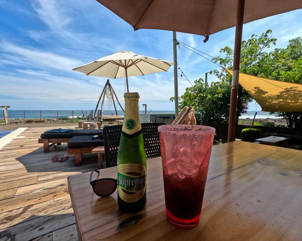 A bottle of “Supreme cerveza” Salvadoran beer next to a glass of ice with a salted rim on a table under an umbrella at a hotel with the Pacific Ocean in the background. ©KettiWilhelm2023