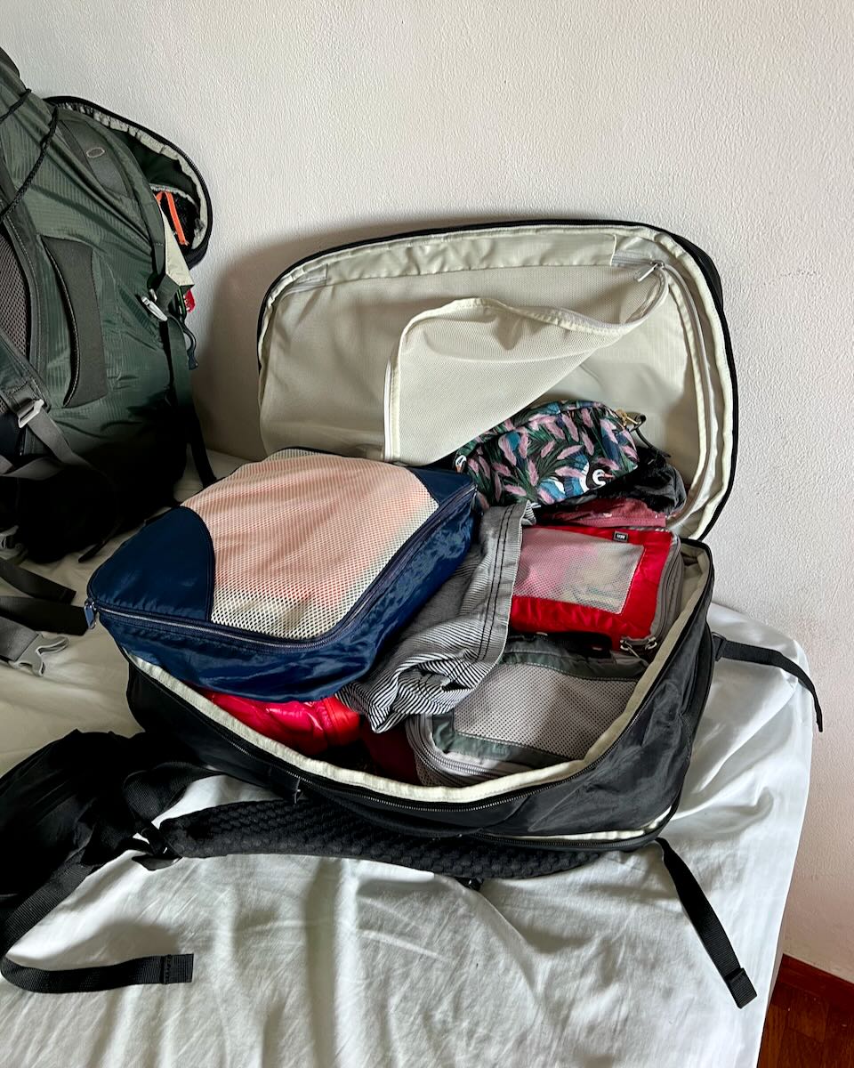 The Tortuga travel backpack on the bed in my hostel, with the clamshell lid open against the wall and items falling out of its zipper compartment. ©KettiWilhelm2023