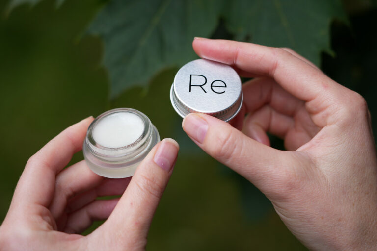 The author of this Rebrand Skincare review holding an open jar of Dewy Balm lip balm, with green leaves in the background, to show the size of the refillable jar – about one inch wide. ©KettiWilhelm2024