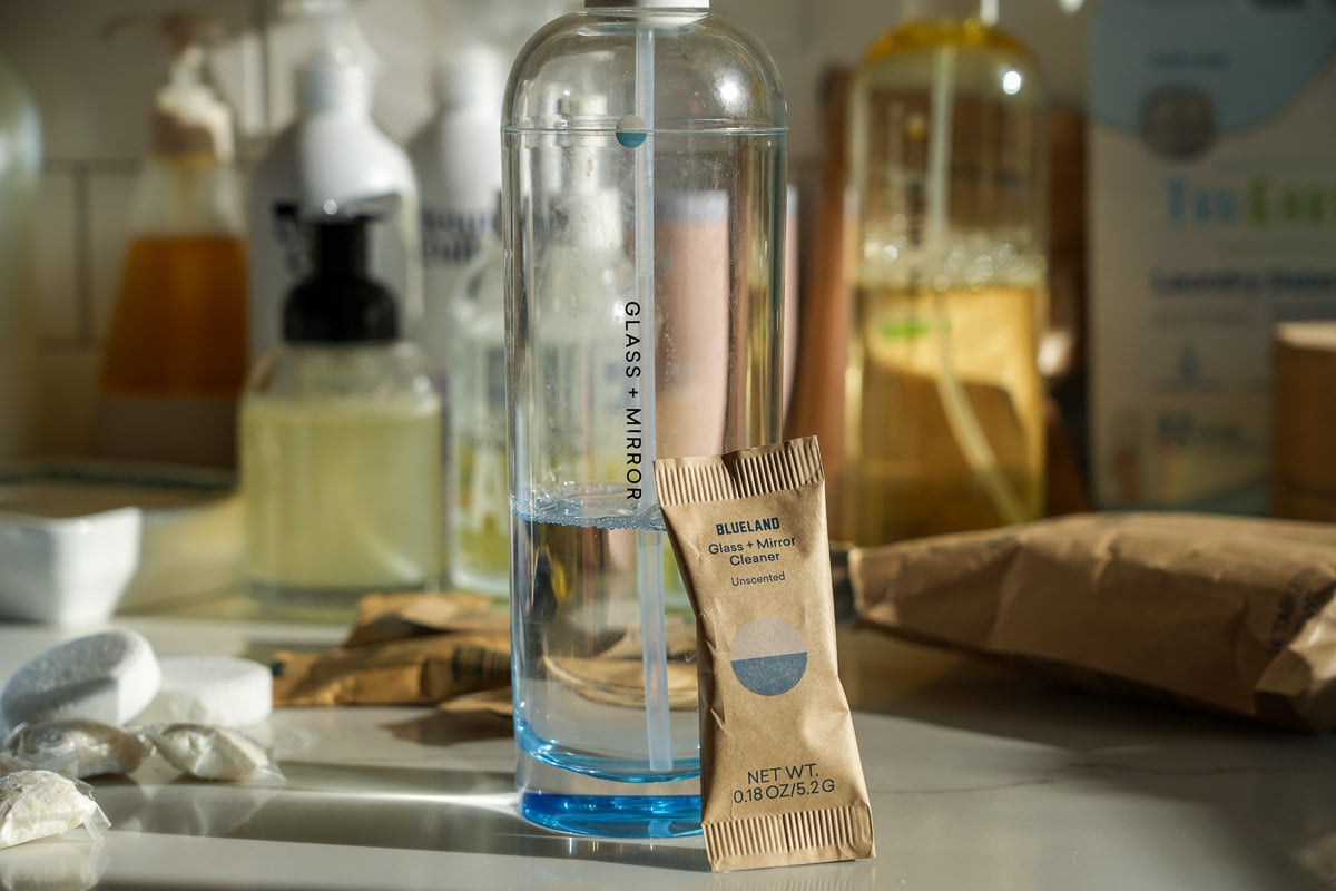 Blueland cleaning tablets for their refillable glass and mirror cleaner, in front of a Blueland bottle from the brand's original design, which was prone to leaking. Blueland's new bottles do not break or leak. ©KettiWilhelm2025