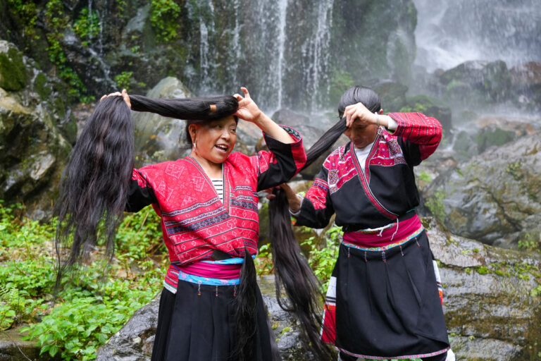 Two Red Yao indigenous women performing the famous Red Yao hair care routine and styling their floor-length hair in their traditional indigenous fashion in front of a waterfall in Longsheng county, Guangxi, rural China. ©KettiWilhelm2025