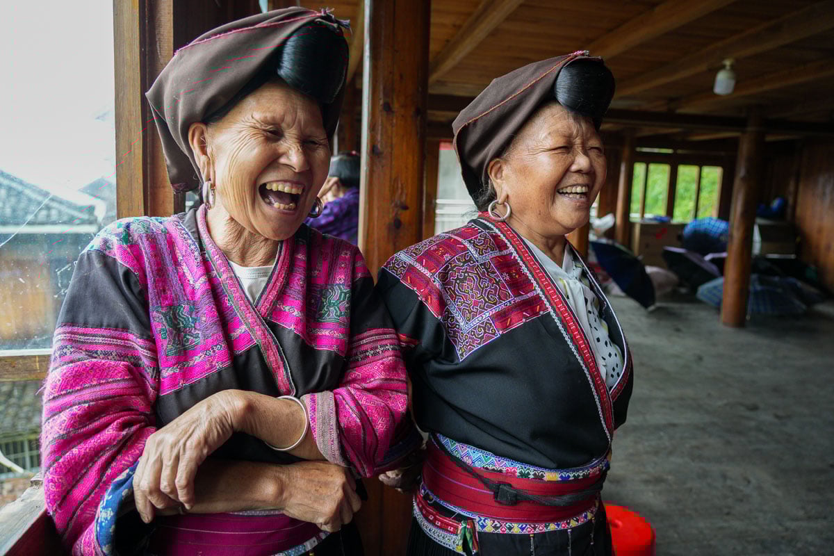 Two Red Yao indigenous women standing next to a window laughing while hosting guests for a community-based travel experience in Longsheng county, Guangxi, rural China. ©KettiWilhelm2025