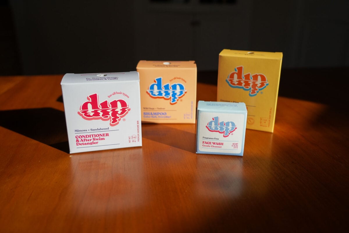 Four plastic-free products from Dip in their colorful, vintage-inspired cardboard boxes, sitting on my wooden table: Shampoo bar, conditioner bar, face wash bar, and body wash bar. ©KettiWilhelm2025