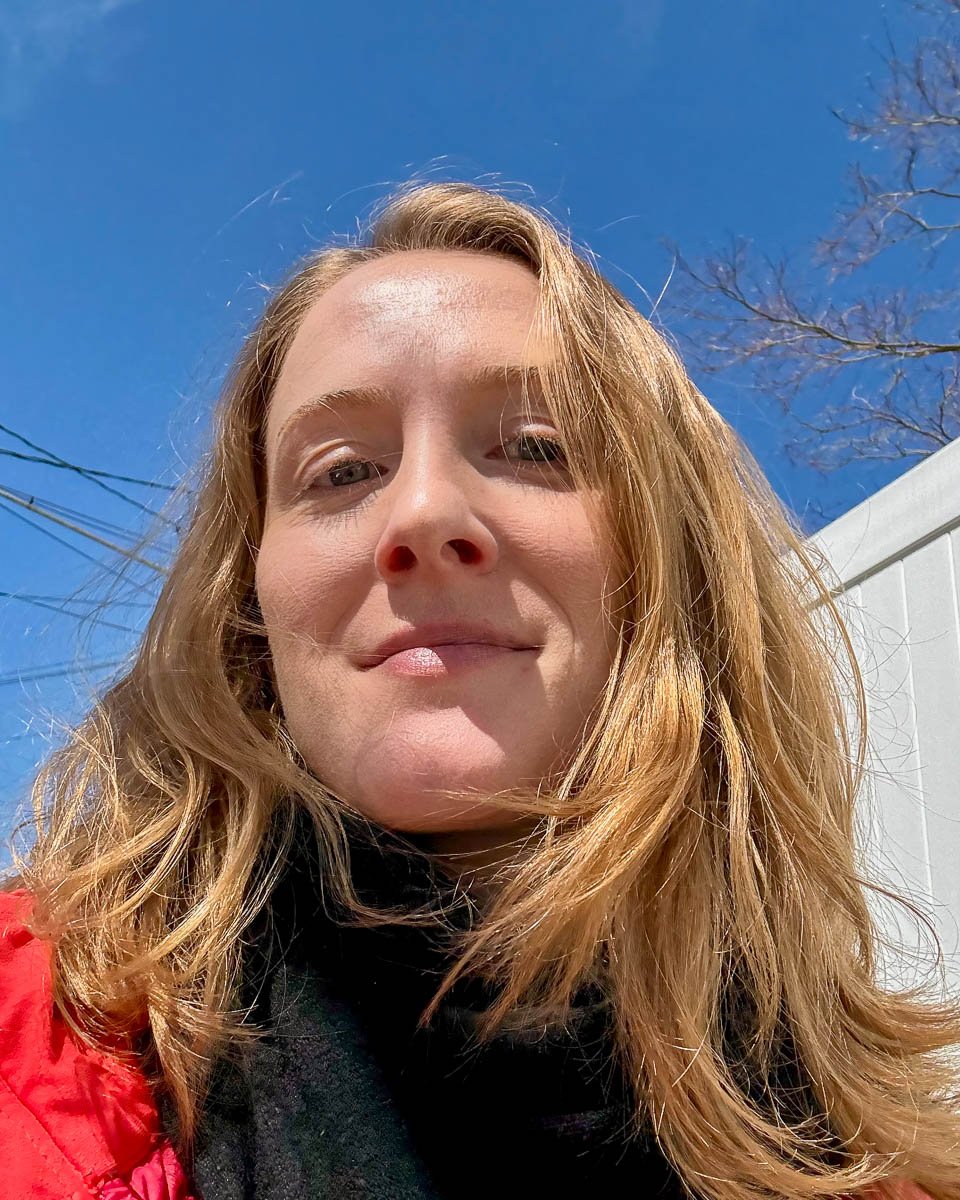 The author of this Avasol sunscreen review, a white woman in her 30s, looking down at the camera under a blue sky with two different shades of Avasol mineral sunscreen stick blended on her face. ©KettiWilhelm2025