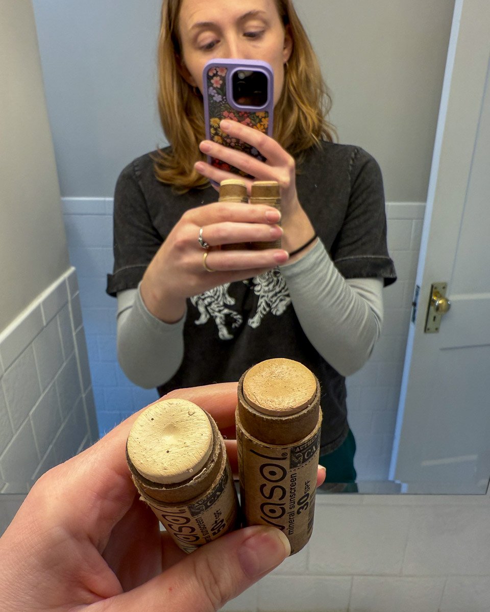 The author of this Avasol sunscreen review, a white woman in her 30s, looking in the mirror holding two different shades of Avasol mineral sunscreen stick – light and medium. ©KettiWilhelm2025