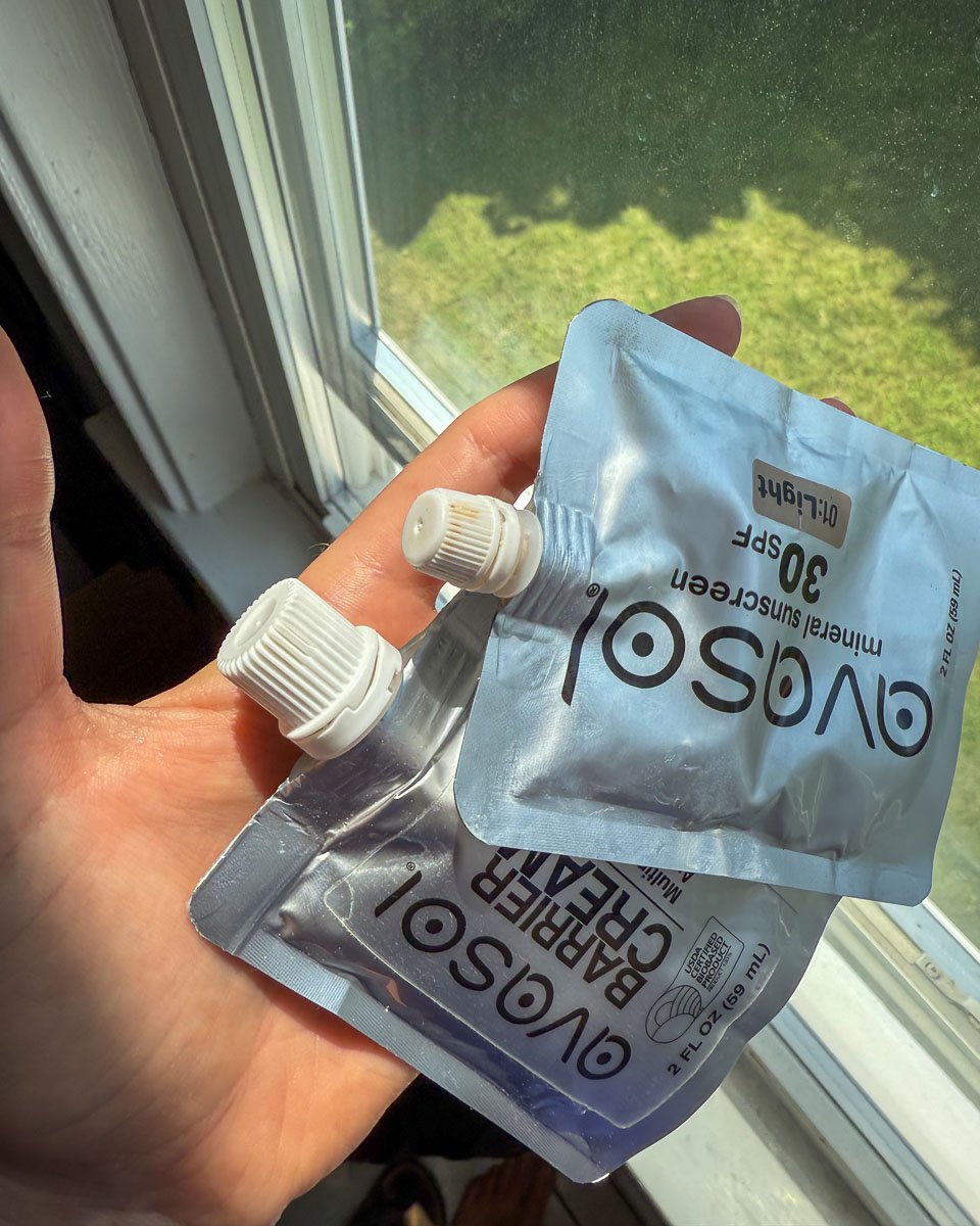 The hand of author of this Avasol sunscreen review, holding the aluminum pouches of their "Environmental Defense Cream" tinted mineral sunscreen cream in the "light" shade and "Barrier Cream" natural, multipurpose skin protectant. ©KettiWilhelm2025