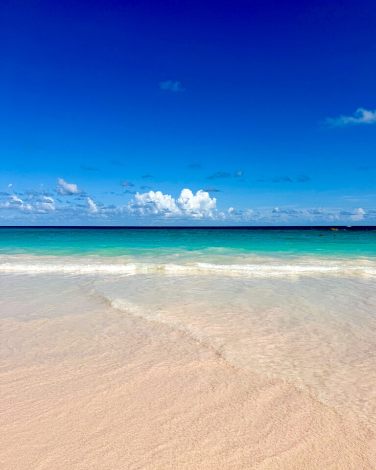 Bright blue sky with only a few white clouds in the distance, turquoise water, and pink sand of Bermuda. ©KettiWilhelm2025
