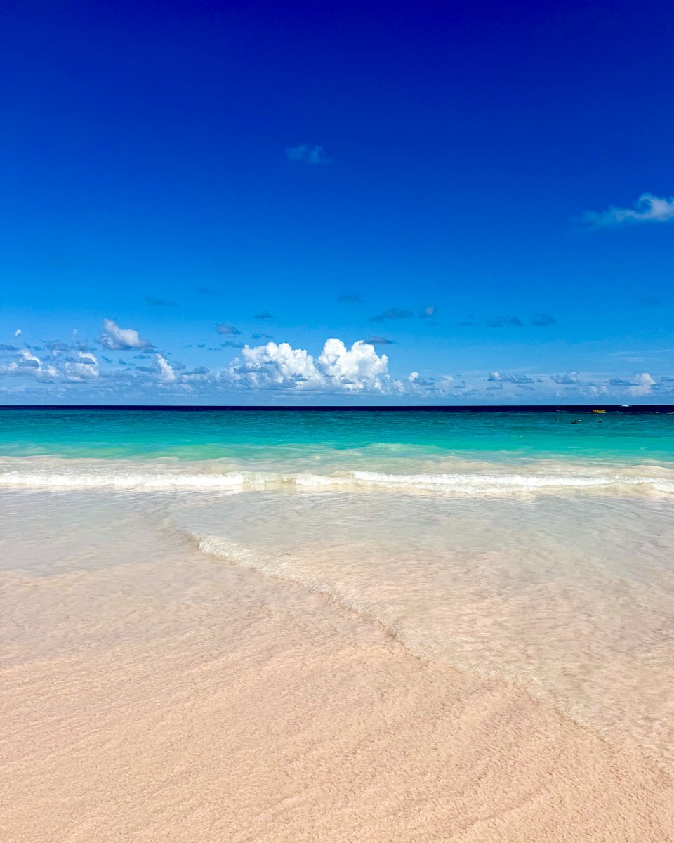 Bright blue sky with only a few white clouds in the distance, turquoise water, and pink sand of Bermuda. ©KettiWilhelm2025