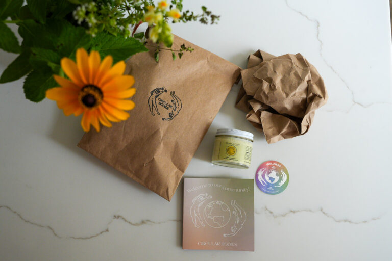 Glass jar of mineral suncream from Circular Bodies, with its compostable and plastic-free shipping materials next to a bouquet of backyard flowers. ©KettiWilhelm2025
