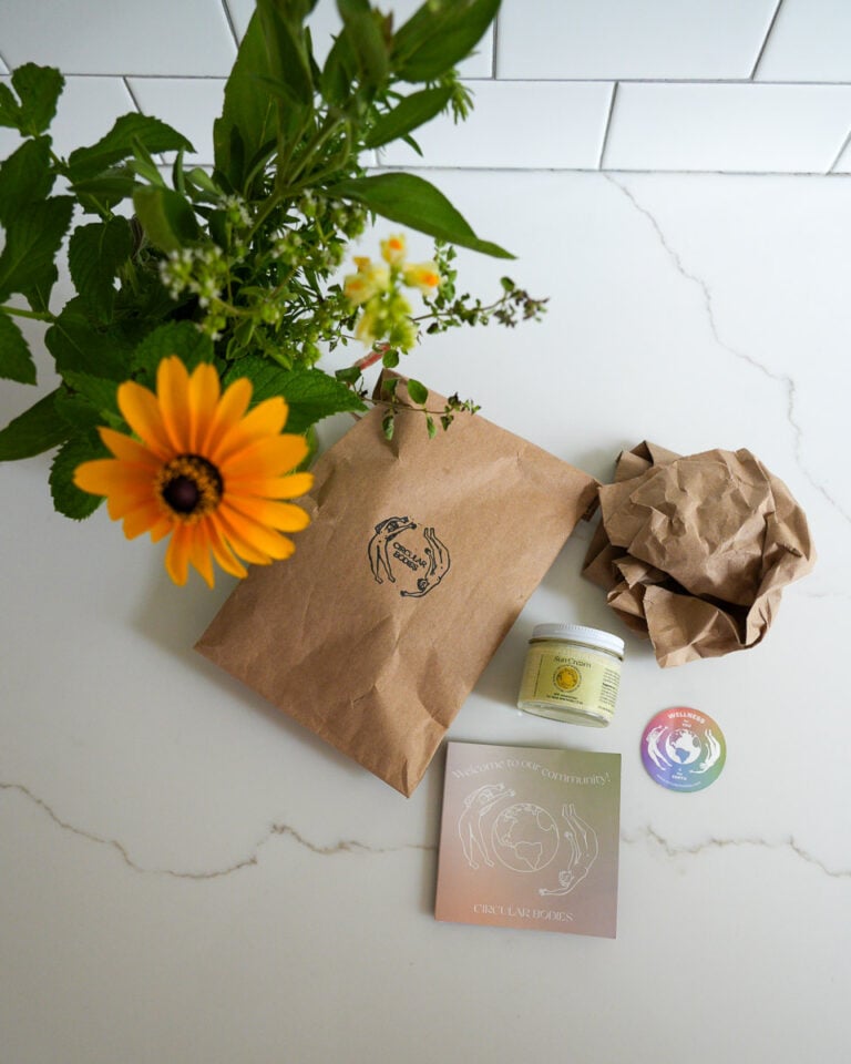 Glass jar of mineral suncream from Circular Bodies, with its compostable and plastic-free shipping materials next to a bouquet of backyard flowers. ©KettiWilhelm2025