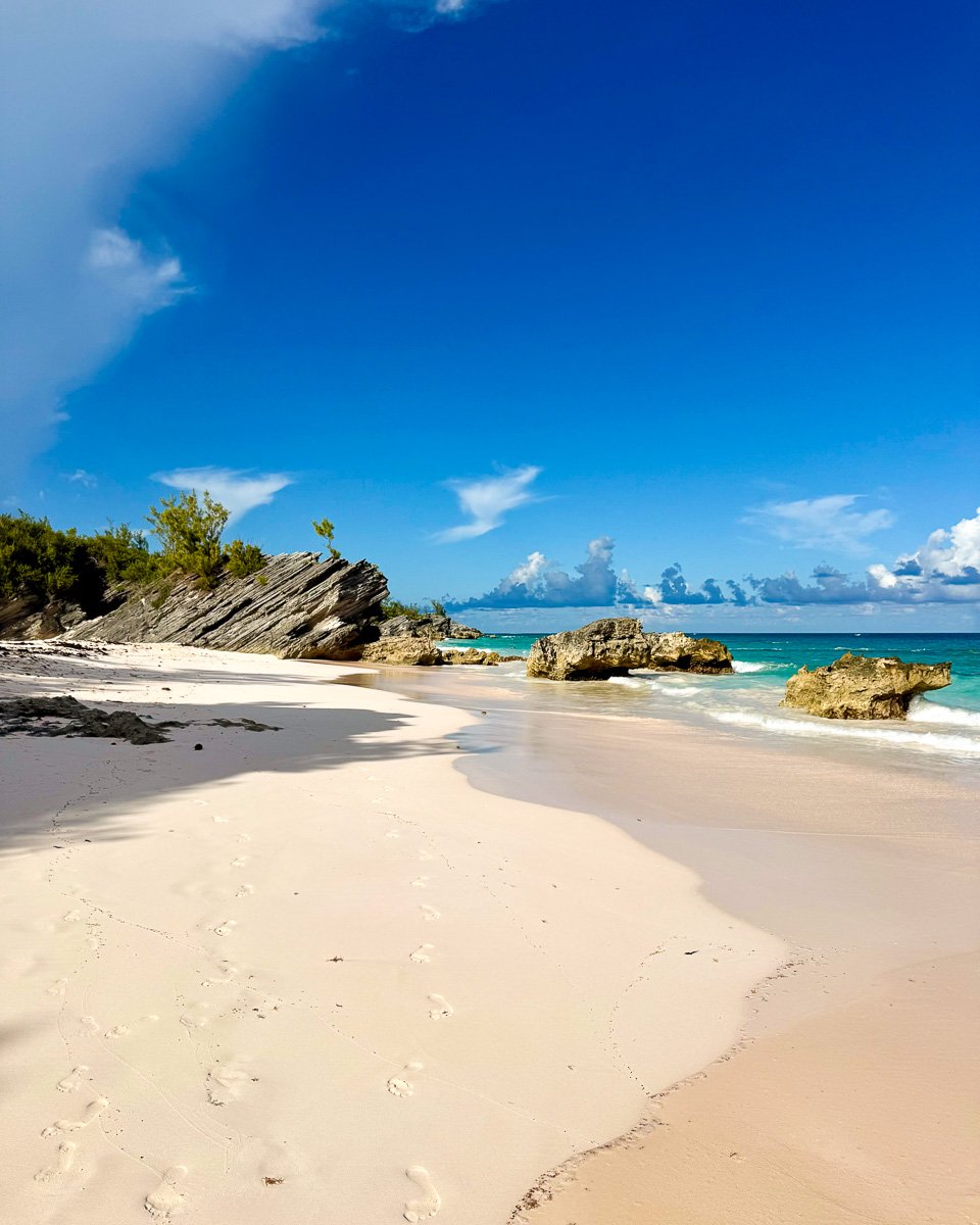Bright blue sky with only a few white clouds in the distance, turquoise water, and pink sand with footprints walking down an empty beach in Bermuda. ©KettiWilhelm2025