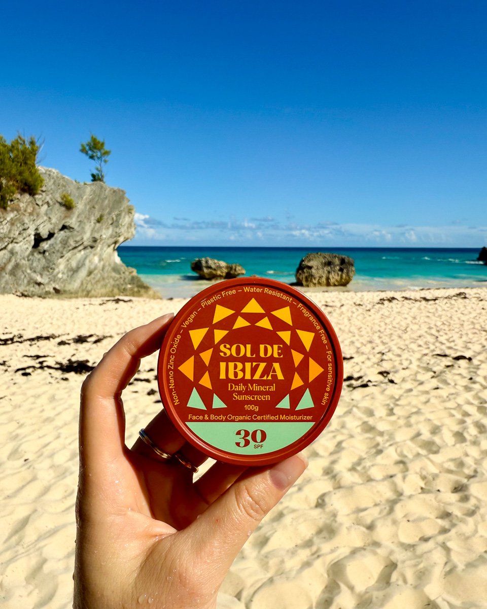 A metal tin of organic sunscreen held in the travel writer's hand in front of a bright blue, cloudless sky over an empty beach in Bermuda. ©KettiWilhelm2025