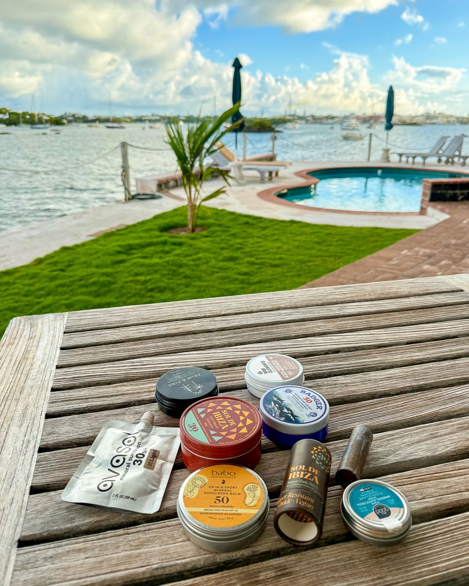 My collection of organic sunscreen sticks on a table in Bermuda, one of the places where I tested these products from several sustainable brands: Avasol, Raw Elements, Palm and Pine, All Good, Badger, Babo Botanicals, and Sol de Ibiza. ©KettiWilhelm2025