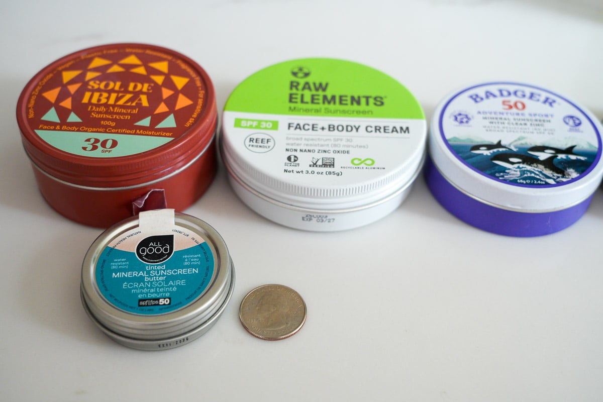 Four metals packages of mineral sunscreen from Sol de Ibiza, Raw Elements, Badger and All Good, next to a quarter to show the small size of All Good's SPF 50 sunscreen. ©KettiWilhelm2026